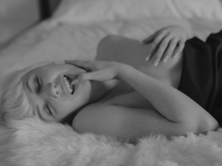 A person with short blonde hair smiles while lying on a bed, partially covered with a blanket. The image is in black and white.