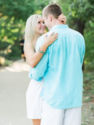 A couple embraces on a sunlit path surrounded by greenery. The woman smiles while looking at the man.