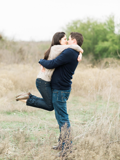 A couple embraces in a field, with the woman lifting her legs as the man holds her. Sparse vegetation is visible in the background.