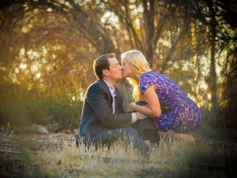 A couple kneels and kisses outdoors amid dry grass and trees, with a soft, warm light illuminating the scene.