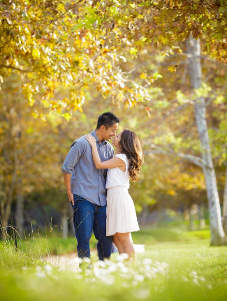 A couple embraces and kisses in a sunlit park surrounded by trees with autumn leaves.