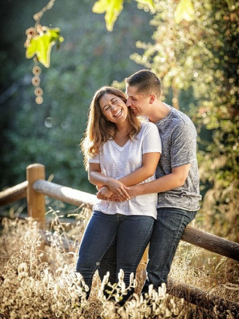 A couple stands outdoors by a wooden fence, smiling and embracing in a sunlit natural setting.