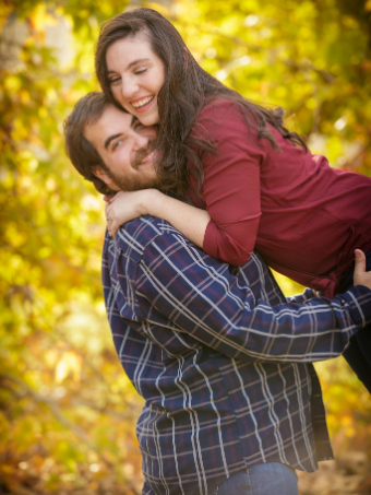 A man in a plaid shirt lifts a smiling woman in a red shirt. They are outdoors in a wooded area with autumn leaves.