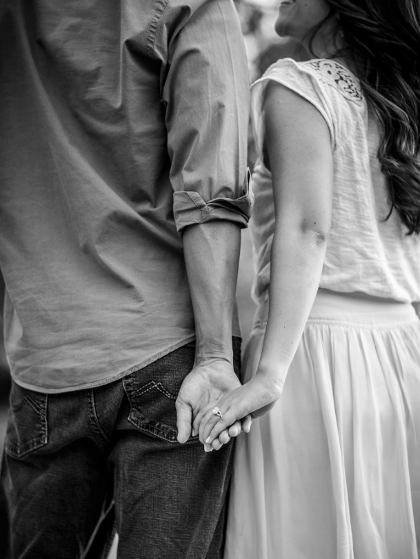 A couple holds hands while walking, with their backs facing the camera. The woman wears a skirt and the man wears jeans. The image is in black and white.