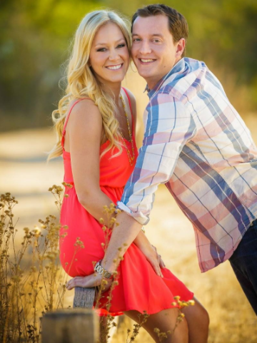 A smiling couple poses outdoors, with the woman in a red dress and the man in a plaid shirt. They lean on a wooden fence surrounded by dry plants.