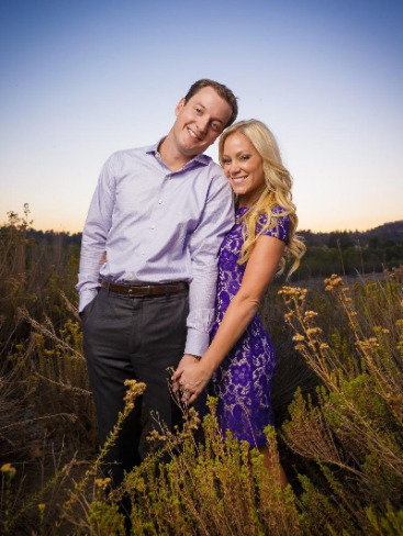 A couple stands close together in a field, holding hands. The man wears a light shirt and dark pants, while the woman is in a purple dress. The sky is clear at sunset.