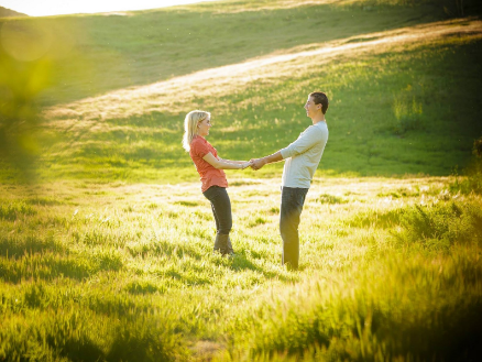 A couple stands in a sunlit grassy field, holding hands and facing each other, with a sloping green hill in the background.