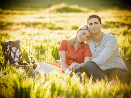 A couple sitting closely on a picnic blanket in a sunlit field, with a picnic basket nearby.