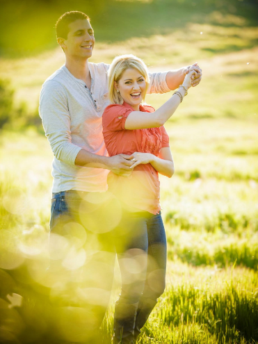 A couple stands in a sunlit field, smiling and holding hands, surrounded by green grass and soft focus foreground.