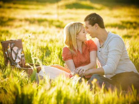 A couple sits on a picnic blanket in a grassy field at sunset, holding hands and looking at each other affectionately, with a picnic basket nearby.