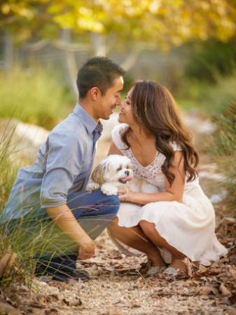 A couple kneels facing each other on a leaf-strewn path, smiling with a small dog between them.