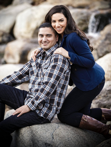 A man in a plaid shirt sits on a rock while a woman in a blue jacket leans on him. They both smile at the camera in an outdoor setting.