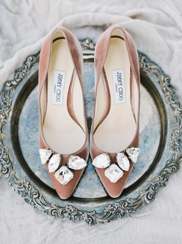 A pair of pink velvet high heels with large crystal embellishments on the toes, placed on an ornate silver platter.