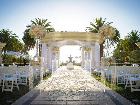 Outdoor wedding aisle lined with white chairs, rose petals, and tall floral arrangements, leading to a decorated altar with drapes under a sunny sky.