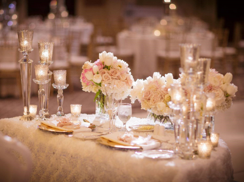 A beautifully set wedding table with elegant glassware, white and pink floral arrangements, and glowing candles on a lace tablecloth.