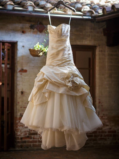 Strapless white wedding dress with layered tulle and fabric flowers, hanging on a hanger in a rustic setting.