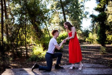 A person kneels and holds the hand of another person standing in a red dress in a forest clearing.