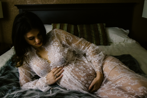 Pregnant woman in a sheer lace gown lies on a bed, gazing at her belly.