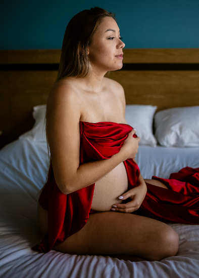 Pregnant woman sitting on a bed, wrapped in red fabric, hands on stomach, looking to the side.