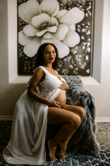 Pregnant woman in a white dress seated on a chair with a large floral painting behind her.