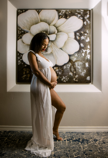 Pregnant woman in a white dress stands in profile, cradling her belly. A large floral painting hangs on the wall behind her.