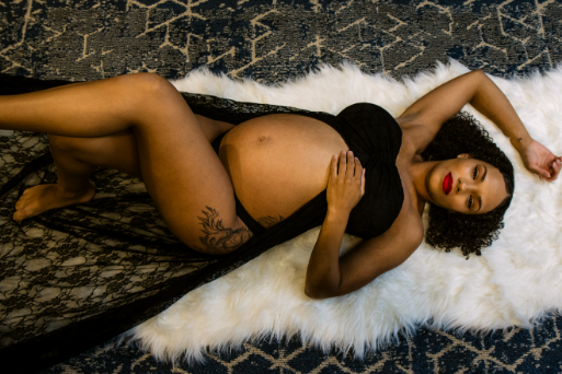 Pregnant person in a black lace outfit lies on a white furry rug on a patterned carpet, with one arm raised and eyes closed.