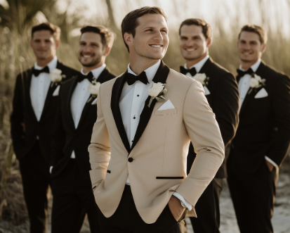 Groom in a beige tuxedo with groomsmen in black tuxedos standing in a line outdoors.